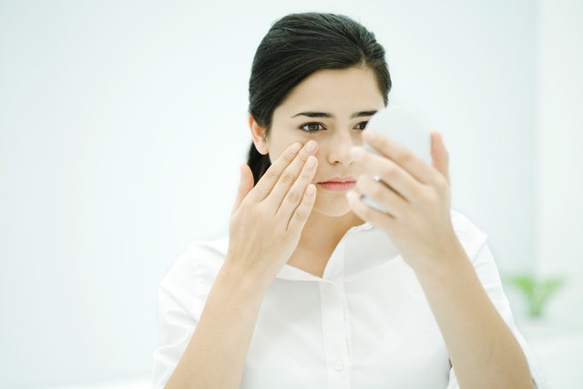 Young woman looking at self in hand mirror, toucing her cheek