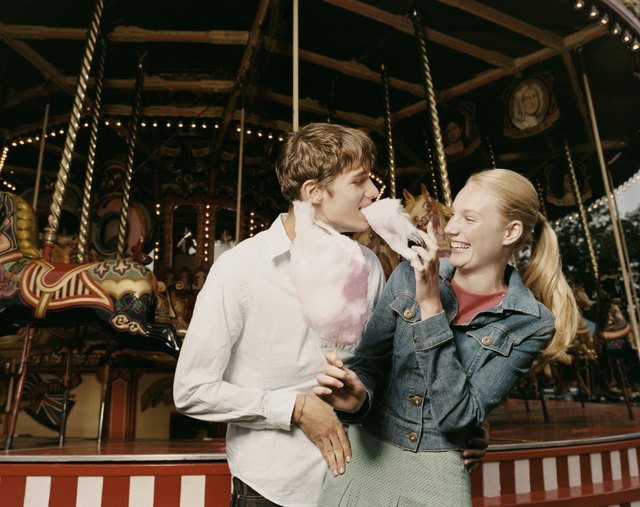Teenage Couple Stand in Front of a Carousel, Girl Feeding Her Boyfriend Candy Floss and Laughing