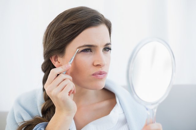 Concentrated casual brown haired woman in white pajamas plucking her eyebrows