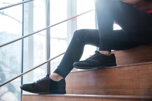 Closeup of legs in black leggings and sneakers of young sportswoman sitting on stairs.