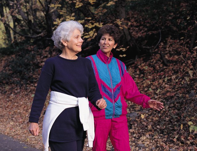 an elderly woman wearing black workout clothes walks with a middle-aged woman wearing red and blue workout clothes on a path covered with dead and fallen leaves