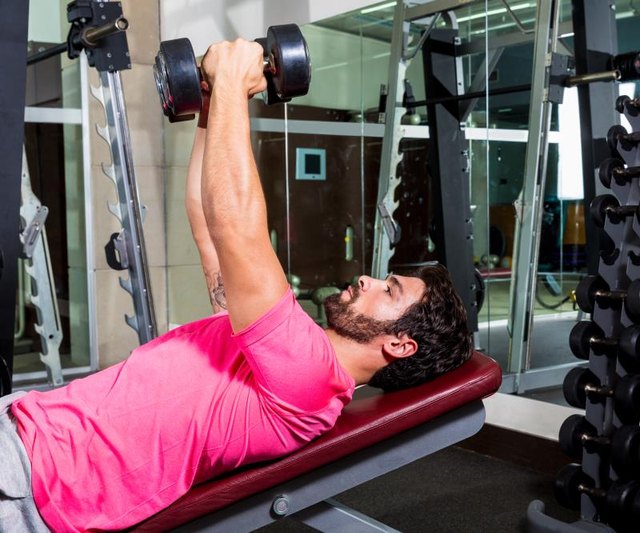 A man with a beard and pink shirt performs inclined dumbbell chest flyes at the gym.