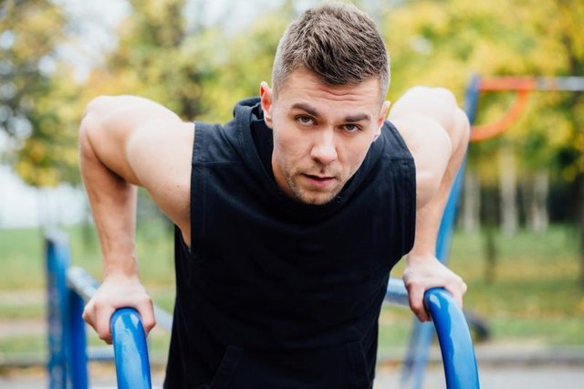A portrait of a focused muscular young man in black workout clothes doing dips on parallel bars at fall city park morning.