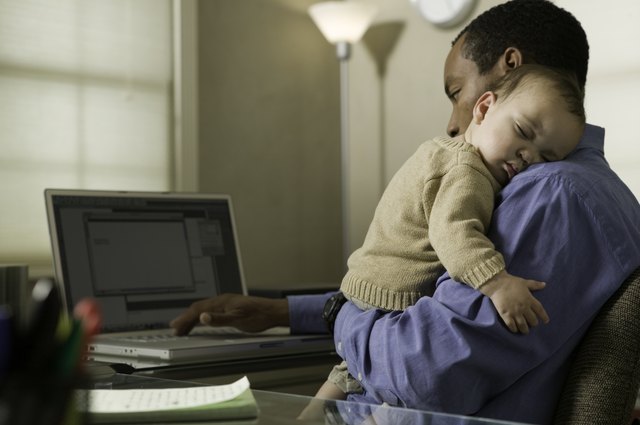 Father using laptop, holding son (9-12 months) on shoulder in home office