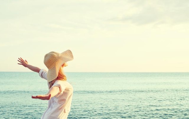 Happy beauty woman in hat is back opened his hands, relaxes and enjoys the sunset over the sea on the beach
