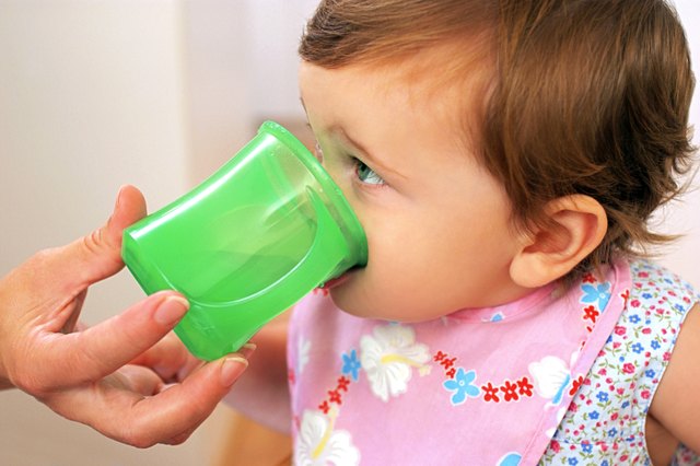 A baby girl drinks from a green cup with help from her mother.