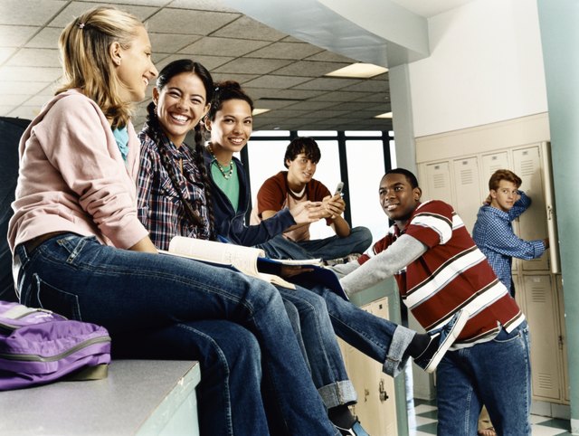Three Female Students Sat on a Row of Lockers, with Three Male Students Looking On