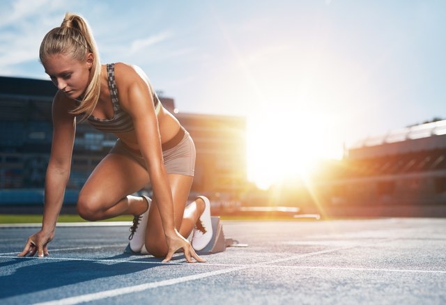 Young woman athlete at starting position ready to start a race. Female sprinter ready for sports exercise on racetrack with sun flare.