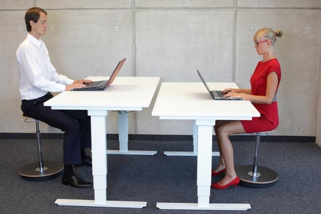 business man and woman coworking in correct sitting posture on pneumatic leaning seats with laptops at electric height adjustable desks in office