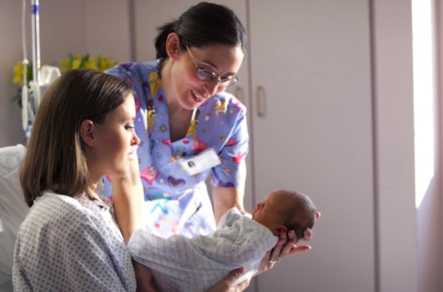 a caucasian mother shows off her newborn baby to a nurse while recovering in a hospital bed