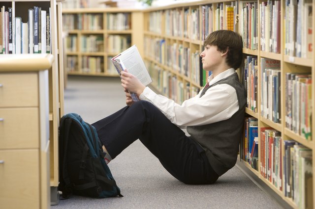 School boy (16-17) reading on floor by bookshelf in library