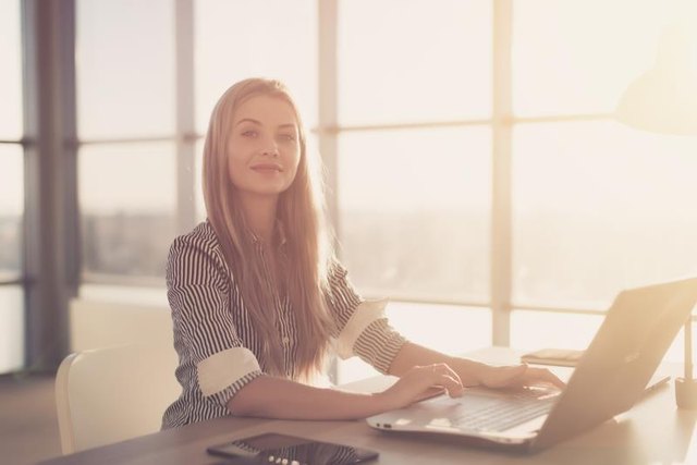 Woman blogging in spacious office using computer on her workplace. Female employee sitting, smiling, looking at camera