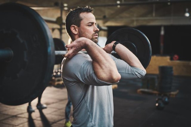 Tough bearded man with thick muscular arms and gray shirt pulling up large barbell in fitness training class indoors