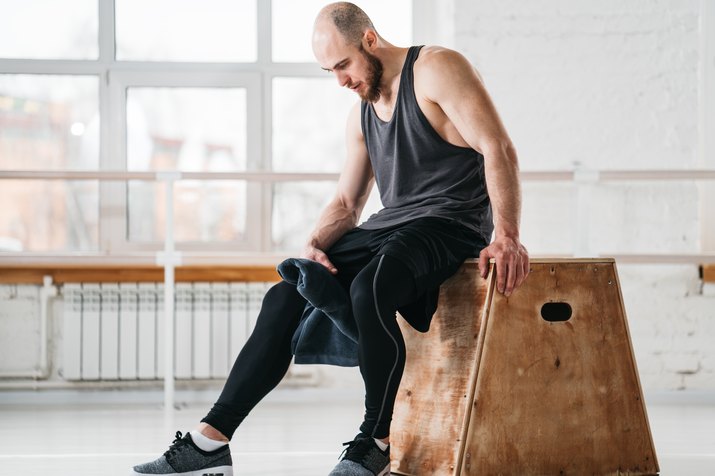 Sweaty muscular man sitting on box in gym gym. Strong male athlete resting after hard workout in light hall