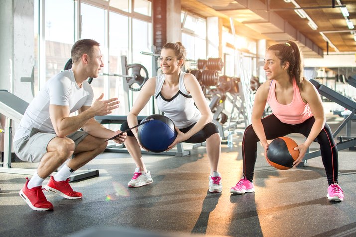 Two sporty women doing exercises with fitness balls with assistance of their personal trainer in gym.