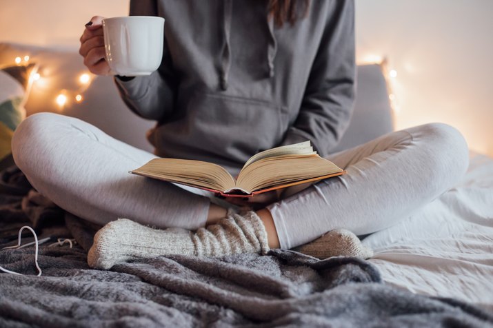 Woman sitting on bed with book and hot drink