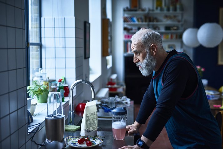 Tired looking senior man leaning on kitchen counter with sports drink