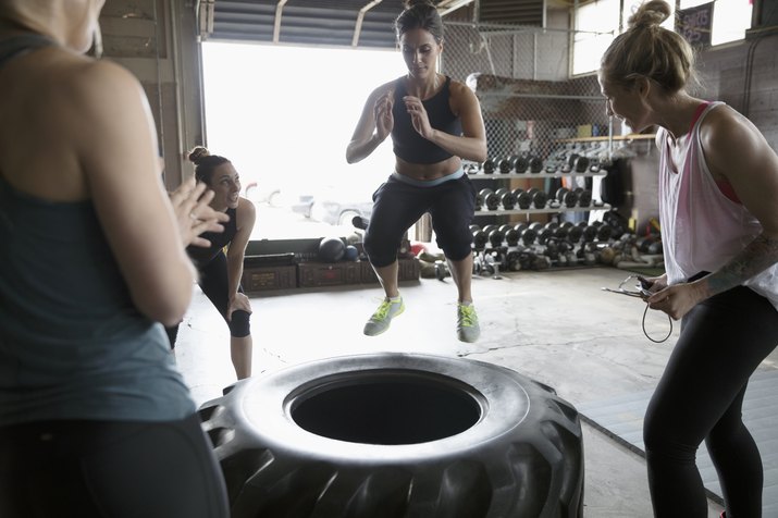 Women cheering classmate jumping on large tire in gym exercise class in gritty gym