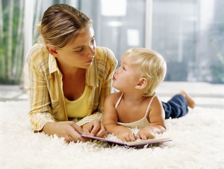 Woman and female toddler (21-24 months) lying on rug with book, looking at each other