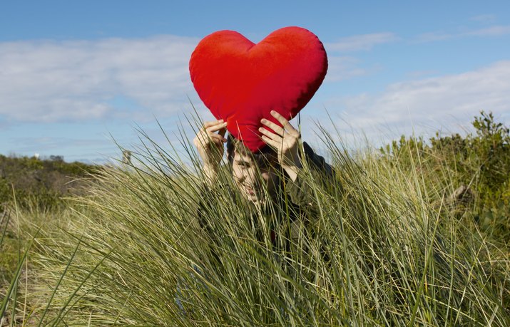 Young man squatting in grass holding up heart-shaped cushion