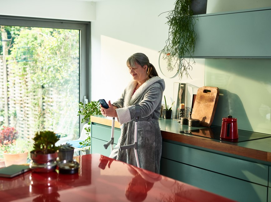 a person with a gray ponytail and forearm crutches wearing a gray bathrobe and standing in their kitchen smiling at their smartphone