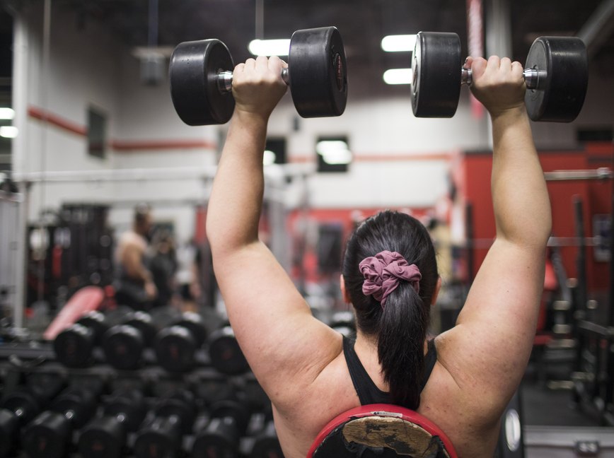 Rear view of a woman wearing a sports bra exercising with dumbbells  at the gym.