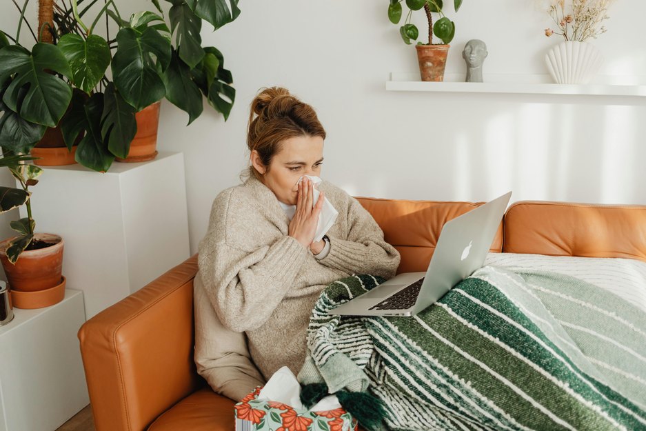 a young adult using nasal congestion home remedies blows their nose on a couch under a blanket with a laptop balanced on their knees