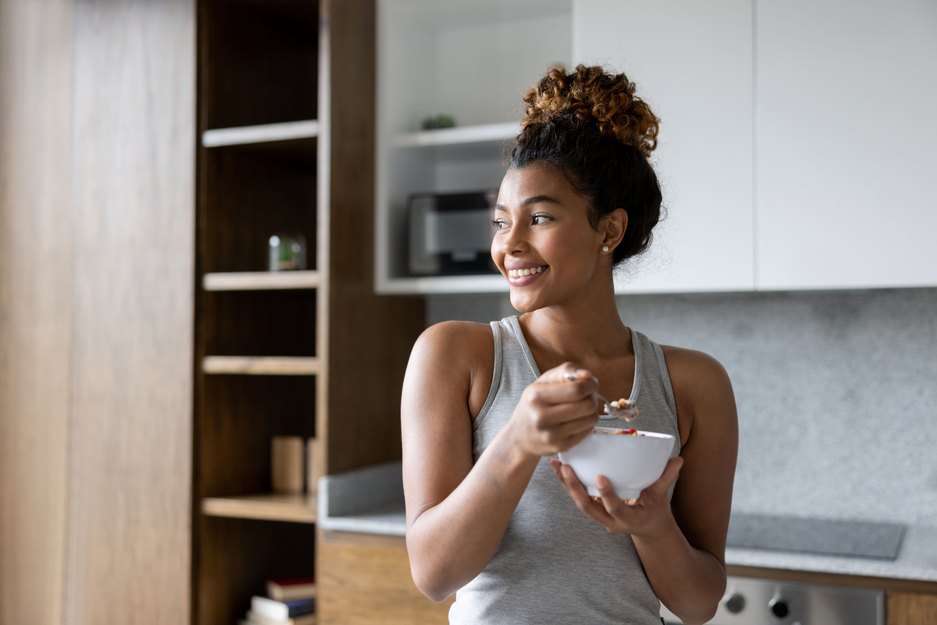 A woman stands in a kitchen smiling and eating from a bowl.