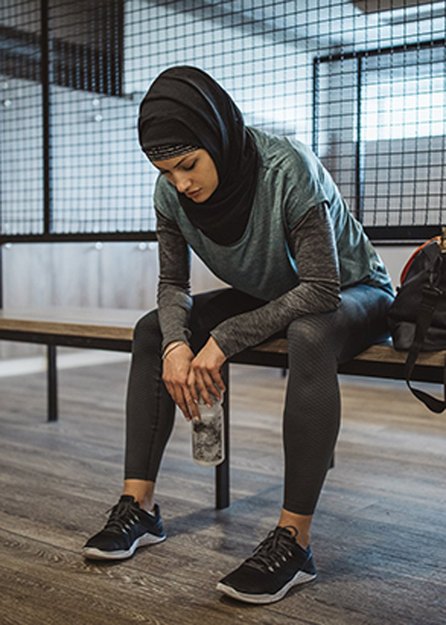a person wearing a gray shirt, black leggings and a black headscarf sits on a bench at the gym looking tired