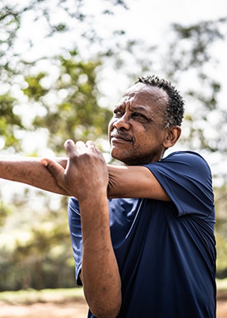 older adult wearing a navy t-shirt doing an arm stretch outside in a park