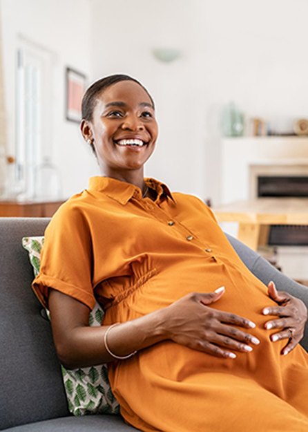 a pregnant person wearing an orange jumpsuit sits on a gray couch holding their stomach and smiling